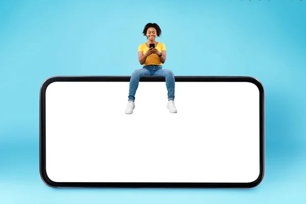 Cheerful young black lady sitting on big smartphone with empty screen, using mobile device, mockup for website — Stockfoto