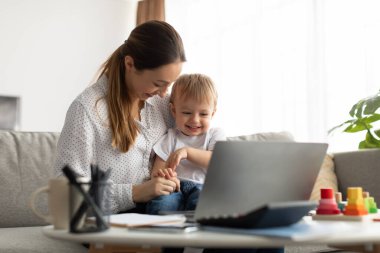 Young mother playing with little son while working on laptop from home, sitting on sofa and tickling child