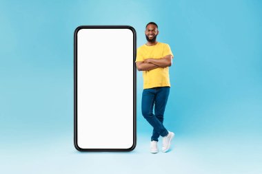 Full length of funky young black guy leaning on huge blank smartphone on blue background, mockup for mobile app