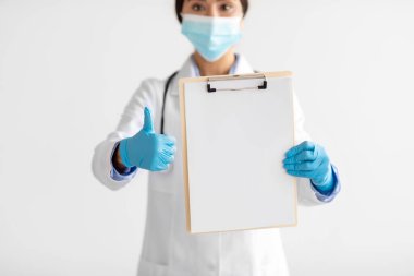 Cropped young cute indian lady doctor in uniform, gloves, protective mask shows tablet with blank space and thumb up