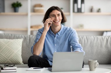 Mobile communication. Happy male freelancer talking on cellphone and working on laptop, sitting on sofa at home