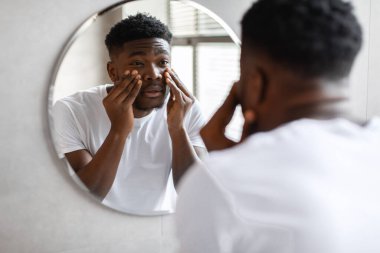 Sleepy black man touching black circles under eyes in bathroom