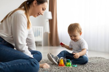 Involved toddler boy with mother playing on floor carpet, using wooden stacking toy, improving fine motor skill of kid