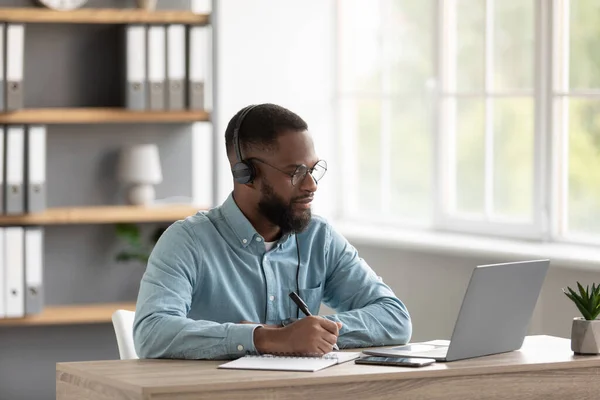 Calm millennial african american male with beard in glasses and headphones watches video lesson on pc — Stockfoto