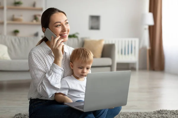 Happy mother with toddler son chatting on smartphone and using laptop computer, sitting on carpet and looking aside — стоковое фото