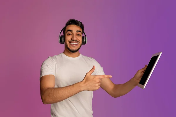 Excited young Arab guy pointing at tablet pc screen, wearing wireless headphones in neon light, mockup — 图库照片