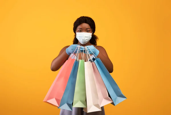 Millennial black woman wearing facial mask and rubber gloves, showing gift bags at camera, making purchases during covid — Stock fotografie
