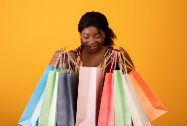 Emotional young black woman looking inside bright shopping bags, satisfied with her purchases over orange background