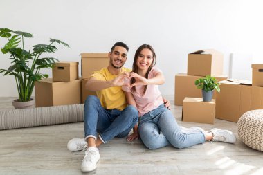 Loving young international couple making heart with their hands, sitting on floor among boxes, moving to new house