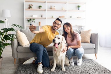 Positive young diverse couple taking selfie with their dog while sitting on floor at home, full length
