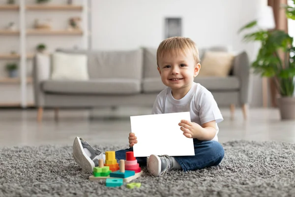 Portrait of adorable little child boy holding blank card and smiling at camera, sitting on floor carpet. Mockup image — Stockfoto