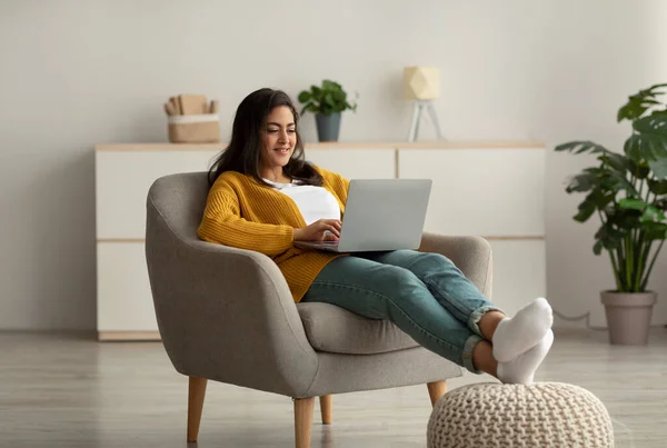 Excited arab woman in casual wear working online from home, sitting in comfy armchair and using laptop, copy space — Foto Stock