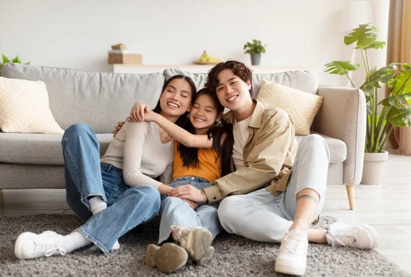 Satisfied happy chinese teen daughter in casual clothes hugs young parents, sitting on floor in living room — Zdjęcie stockowe