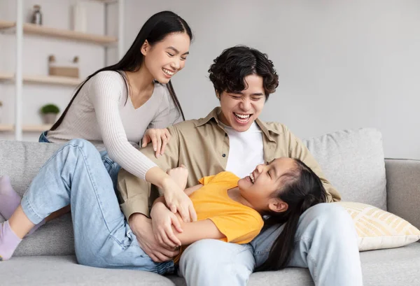 Happy teenager girl and millennial asian man and woman have fun together, tickling little daughter on sofa — Stockfoto