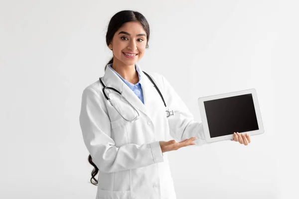 Satisfied young cute indian woman nurse in uniform shows digital tablet with blank screen — Stok fotoğraf