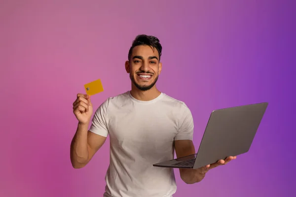 Portrait of cheery millennial Arab man holding credit card and laptop in neon light. Online shopping, remote banking — Zdjęcie stockowe