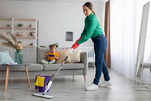 Молода мати прибирає будинок, витирає підлогу і прибирає, поки маленький син дивиться мультфільм на смартфон — стокове фото