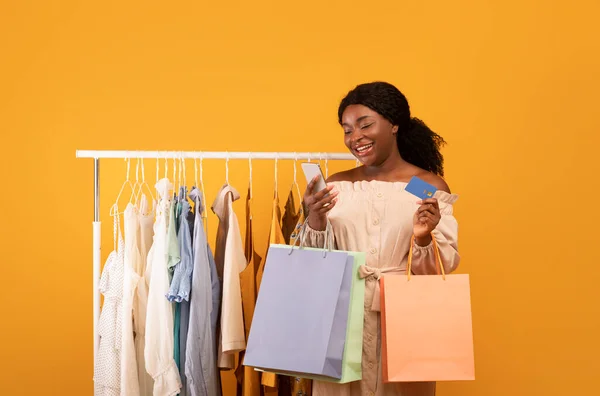 オンラインショッピングのためのスマートフォンやクレジットカードを使用して黒の女性,買い物袋を保持します,衣類ラックの近くに立って — ストック写真