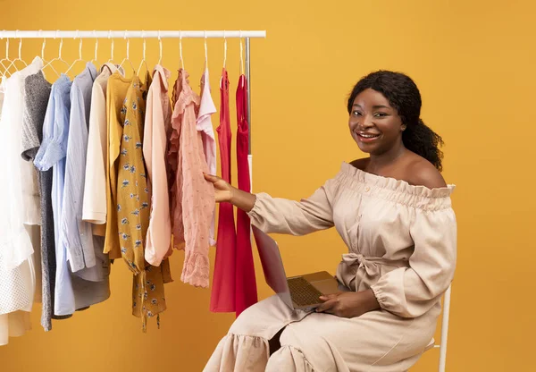 Young black woman with laptop touching clothes on rack, shopping online for new outfits, purchasing on internet — Stockfoto