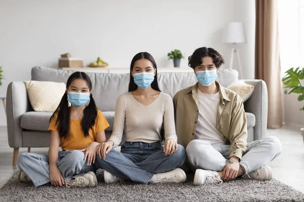 Cheerful teenage girl and young chinese parents in protective masks sitting on floor in living room interior — Stockfoto