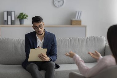 Arab male psychologist talking with female client and taking notes during psychotherapy session, sitting in office