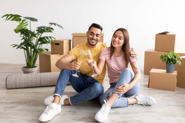Millennial multiracial couple having housewarming party, sitting on floor with champagne, celebrating new home purchase