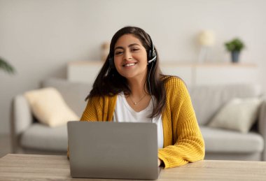 E-learning concept. Happy arab woman wearing headset and studying online with laptop at home, sitting at desk