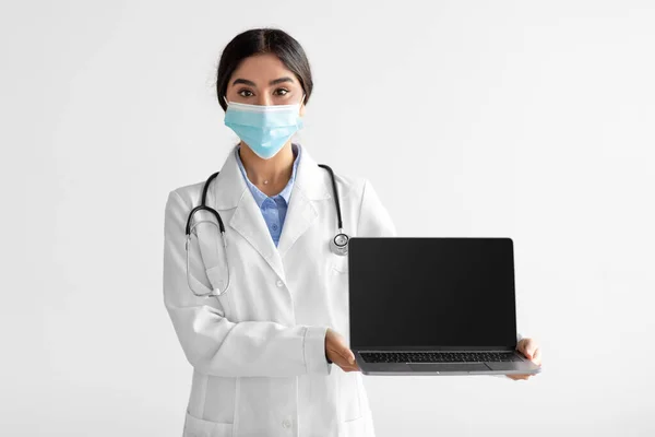 Young attractive hindu woman doctor in uniform and protective mask shows computer with blank screen — Foto Stock