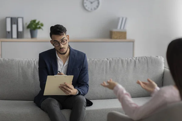 Arab male psychologist talking with female client and taking notes during psychotherapy session, sitting in office — Stockfoto