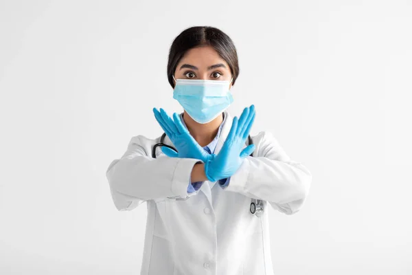 Serious young indian lady doctor in uniform, gloves and protective mask show sign with hands, say no, cross arms — Stockfoto