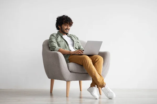 Happy indian guy in casual sitting in armchair with laptop — стоковое фото
