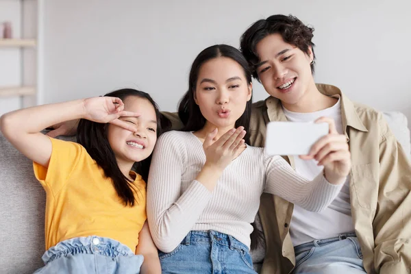 Cheerful young asian guy hugs wife, lady makes blowing kiss, teen daughter makes peace sign and take photo — Stockfoto