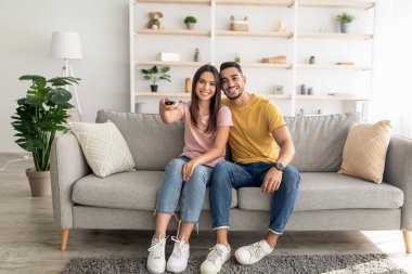 Joyful diverse couple watching TV program, sitting on couch at home, full length portrait