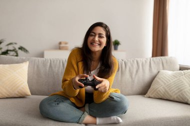 Happy middle eastern lady holding joystick, playing videogames while resting on couch and having fun with computer game