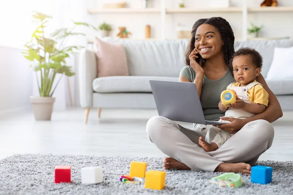 Remote Work With Baby. Black Mom Using Laptop And Talking On Cellphone — стоковое фото