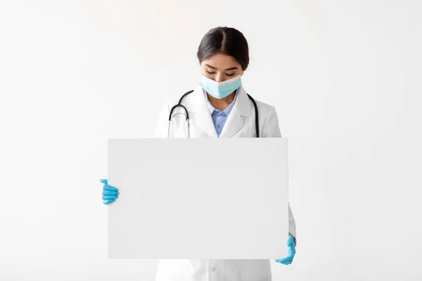 Millennial hindu female nurse in uniform, gloves and protective mask looks at banner with empty space — Stock fotografie