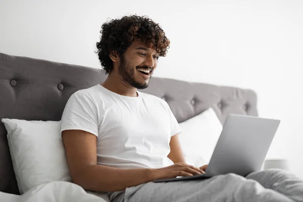 Young indian man typing on laptop, working in bed — Stockfoto