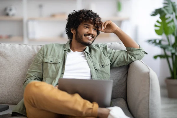 Happy indian guy in casual sitting on couch with laptop — стоковое фото