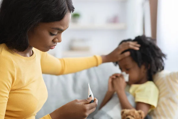 Worried Black Mom Checking Temperature Of Sick Little Daughter Lying On Couch — стоковое фото