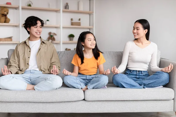 Glad happy pretty teen korean girl, millennial mother and father sits in lotus position, practice yoga on sofa — Stockfoto