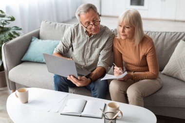 Senior Couple Using Laptop Holding Paper Taking Notes At Home