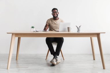 Smiling African American man writing in notebook, taking notes and using computer, sitting at desk in home office, copy space