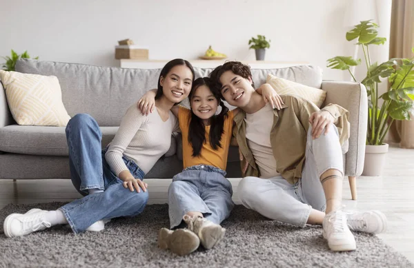 Satisfied happy young asian mother, father and teen daughter hugs, sits on floor in living room interior