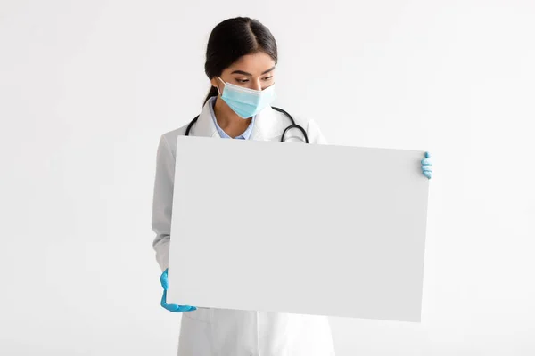 Young indian woman doctor in uniform, gloves and protective mask shows banner with blank space —  Fotos de Stock
