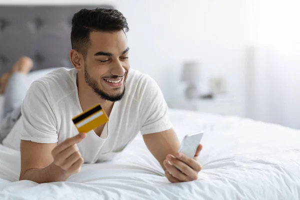 Online Shopping. Happy Arab Guy Using Smartphone And Credit Card In Bed — Zdjęcie stockowe