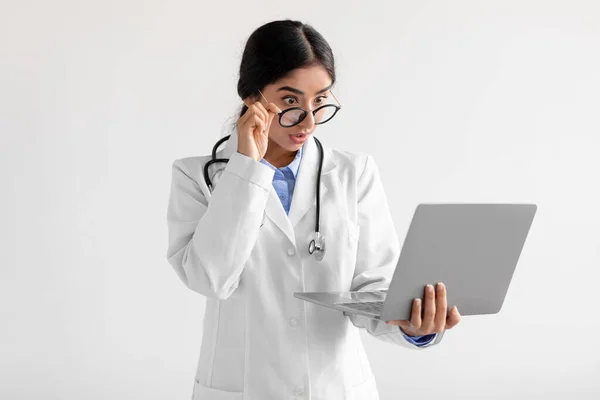 Surprised young cute indian lady medical worker in uniform takes off glasses and looks at computer — Foto Stock