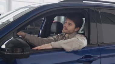 Young dreamy guy sitting in new car at dealership salon, enjoying his purchase and showing thumb up to camera, zoom in