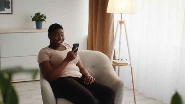 Emocionado mujer afroamericana leyendo mensaje en el teléfono inteligente en casa — Vídeo de stock