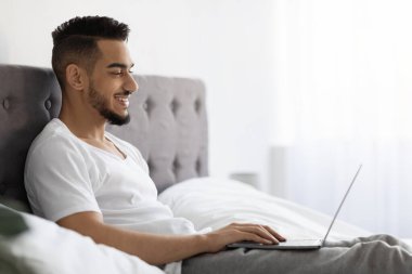 Smiling Young Arab Guy Using Laptop While Relaxing In Bed At Home