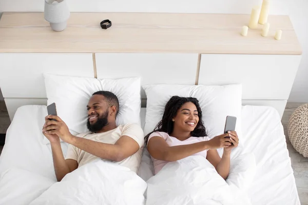 Satisfied handsome young black wife and husband typing on phones, chatting, playing online, lying on bed — Stock fotografie